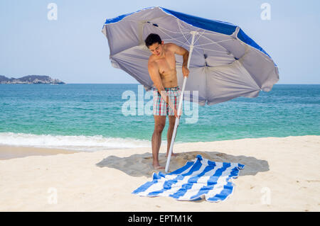 Athletische Männchen setzen einen blauen und grauen Schirm am Strand neben einem Handtuch mit dem Ozean im Hintergrund. Stockfoto