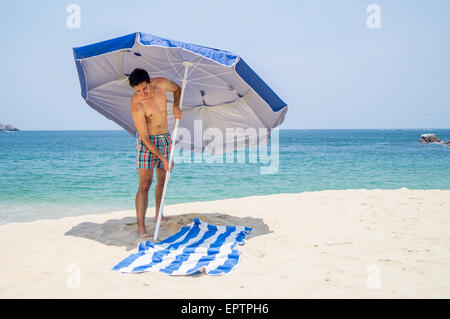 Athletische Männchen setzen einen blauen und grauen Schirm am Strand neben einem Handtuch mit dem Ozean im Hintergrund. Stockfoto