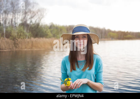 Sommersprossige Mädchen mit Hut und Blumenstrauß steht am Ufer des Sees Stockfoto