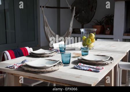 Patio Tisch für das Mittagessen mit Rohrstock underplates, Qhite Platten, Türkis Brille, Zitronen auf Holztisch mit Hängesessel im Hintergrund in südafrikanischen Strandhaus Stockfoto