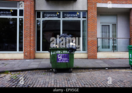 Eine grüne Metall Wheelie bin überfüllt mit schwarzen Mülltüten auf einer gepflasterten Straße in Bristol. Stockfoto