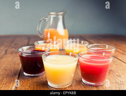 sechs Gläser verschiedener Bio-Säfte mit einem Krug Saft im Hintergrund auf einem Holztisch vintage Stockfoto