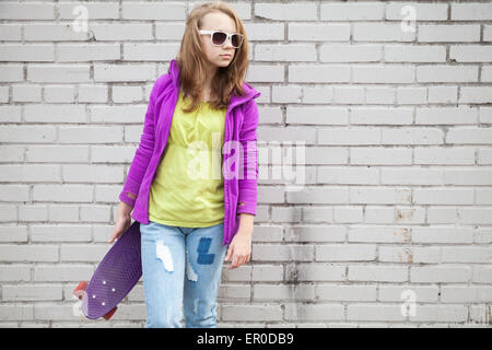 Blonde Teenager-Mädchen in Jeans und eine Sonnenbrille hält Skateboard in der Nähe von grau urban Mauer Stockfoto