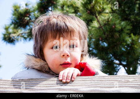 Kopf- und Schulteraufnahme des kleinen Kindes, Jungen, 3-4 Jahre alt, mit roten Wangen, im Wintermantel mit pelzigen Kragen, blickt über einen Holzzaun auf den Betrachter. Stockfoto