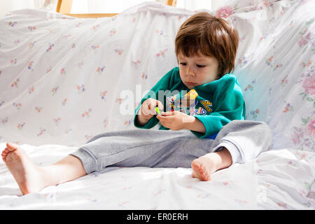 Kind, Junge, 3-4 Jahre alt, auf einem Sofa liegend, während mit einem kleinen Spielzeug, das er mit beiden Händen hält, konzentrierte Ausdruck. Stockfoto