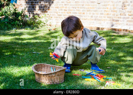 Kind, Junge, 4-5 Jahre alt, in hellem Sonnenlicht, hocken auf dem Rasen im Garten, während die Kleider-Heringe und legte sie in einen Weidenkorb. Stockfoto
