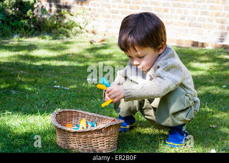 Kind, Junge, 4-5 Jahre alt, in hellem Sonnenlicht, hocken auf dem Rasen im Garten, während die Kleider-Heringe und legte sie in einen Weidenkorb. Stockfoto