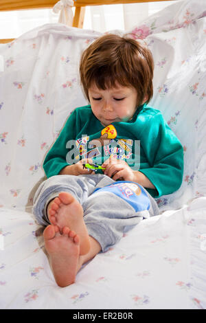 Kind, Junge, 3-4 Jahre alt, auf einem Sofa liegend, während mit einem kleinen Spielzeug, das er mit beiden Händen hält, konzentrierte Ausdruck. Stockfoto