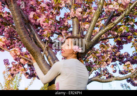 Junges Mädchen mit Blumen im Haar umarmt Baum in Blüte Stockfoto