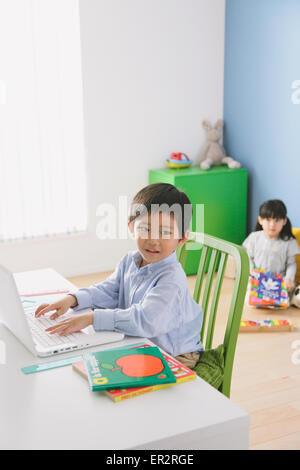 Grundschulkind junge seine Hausaufgaben an seinem Schreibtisch mit seiner Schwester auf dem Boden im Hintergrund lesen Stockfoto