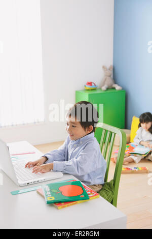 Grundschulkind junge seine Hausaufgaben an seinem Schreibtisch mit seiner Schwester auf dem Boden im Hintergrund lesen Stockfoto