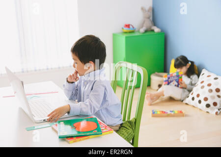 Grundschulkind junge seine Hausaufgaben an seinem Schreibtisch mit seiner Schwester auf dem Boden im Hintergrund lesen Stockfoto