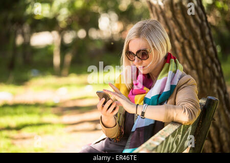 Junge modebewusste Frau nutzt Smartphone auf Bank im Park sitzen. Stockfoto