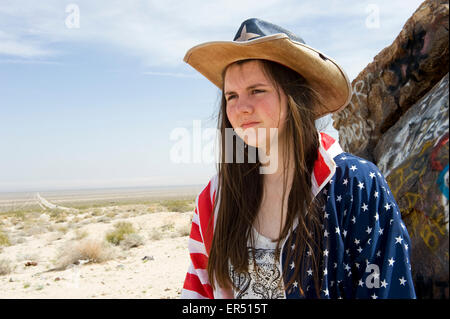 Mädchen in Sternen und Streifen Jacke und Hut stand neben Graffiti bedeckt Rock mit Straße in Ferne, Mojave-Wüste in Kalifornien Stockfoto