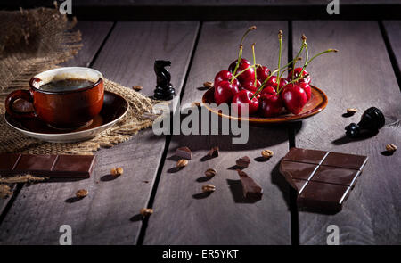 Kaffeetasse, dunkle Schokolade und Kirschen auf dem Holztisch mit schwarzen Schachfigur in der Nähe Stockfoto