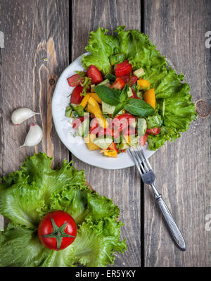 Frischer grüner Salat auf dem Teller, Tomaten auf grünen Blättern und Knoblauch auf dem Holztisch in der Küche Stockfoto