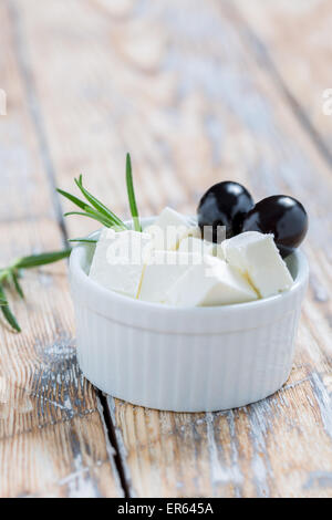 Feta-Scheiben mit Oliven und Rosmarin in einer weißen Schüssel auf einem rustikalen Holztisch Stockfoto