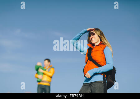Eine junge Frau beschattet Augen gegen Sonnenuntergang mit Baby und Mann im Hintergrund von einer Winterwanderung. Stockfoto