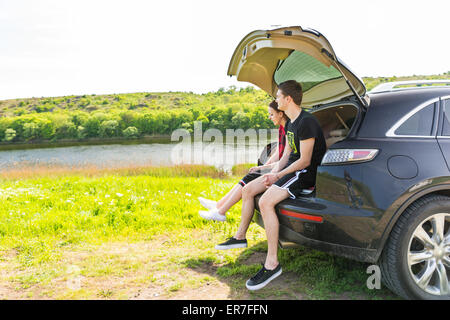 Junges Paar sitzt auf Heckklappe Heckschürze des SUV-Fahrzeug geparkt im grünen Feld Fluss an sonnigen Tag, die Aussicht genießen Stockfoto