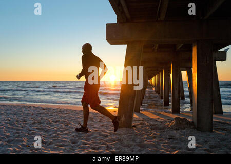 Mann läuft unter einem Steg am Strand bei Sonnenuntergang Stockfoto