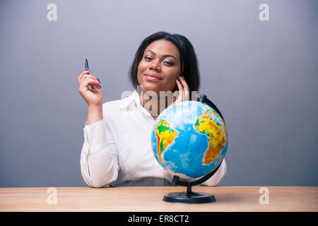 Afrikanische Geschäftsfrau sitzen am Tisch mit Feder und Kugel über grauen Hintergrund und Blick in die Kamera Lächeln Stockfoto