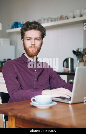 Porträt von zuversichtlich junger Mann mit Laptop am Tisch in der Küche Stockfoto