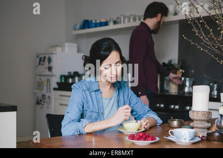 Junge Frau unter Rühren Kaffee am Küchentisch mit Mann im Hintergrund Stockfoto