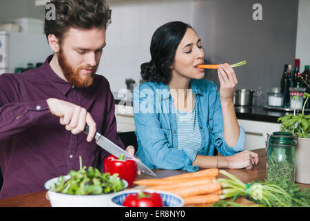 Junger Mann hacken rote Paprika neben Frau Essen Karotte am Küchentisch Stockfoto
