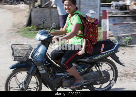 Eine Frau auf einem Motorrad lächelt in die Kamera, als sie vorbei reitet. Ihr Kind ist auf dem Rücken in einem Tuch Riemen geschleudert. Stockfoto