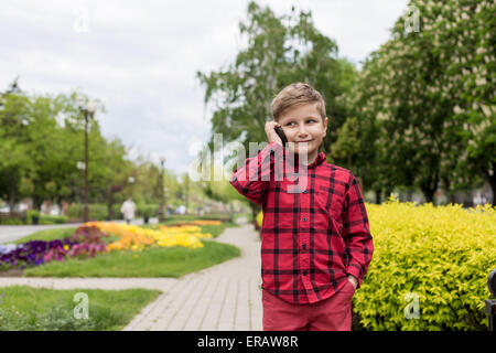 kleiner Junge im roten Hemd am Handy im freien sprechen. Junge lächelt Stockfoto