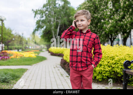 kleiner Junge im roten Hemd am Handy im freien sprechen. Junge lächelt Stockfoto