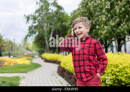 kleiner Junge im roten Hemd am Handy im freien sprechen. Junge lächelt Stockfoto