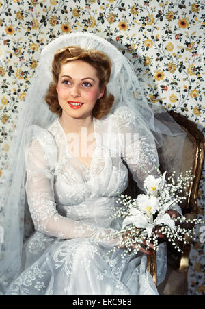 1940ER JAHRE PORTRAIT LÄCHELND BLONDE FRAU BRAUT SITZEN IM STUHL TRÄGT HOCHZEITSKLEID UND SCHLEIER HOLDING BLUMENSTRAUß Stockfoto