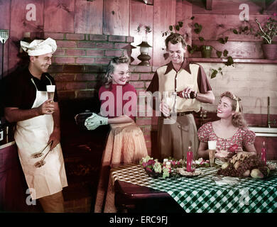 DER 1950ER JAHRE ZWEI PAARE MÄNNER FRAUEN IN KÜCHE TRINKEN BIER ESSEN BURGER MANN TRÄGT KÜCHENCHEF HUT SCHÜRZE VON GEMAUERTEN KAMIN STEHEND Stockfoto