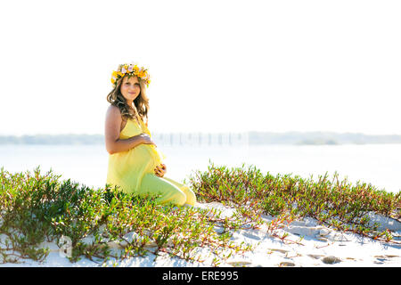 Junge und schöne lächelnd schwangere Frau mit Blüten sitzen an einem Strand Stockfoto