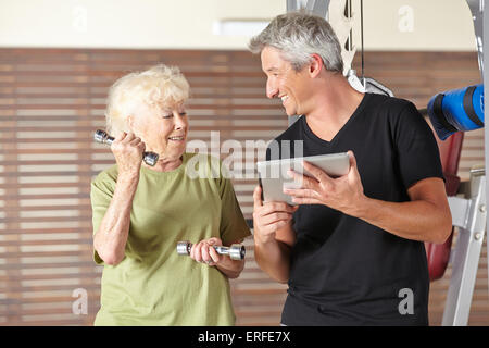 Ältere Frau Training mit Hanteln im Fitnessstudio und Ausbilder mit Tablet-PC Stockfoto