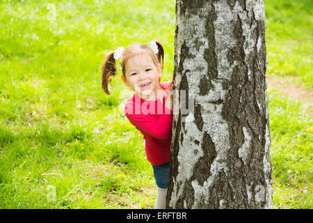 niedliche kleine Mädchen versteckt sich hinter riesigen Baum. Kinder Porträt Blick in die Kamera Stockfoto
