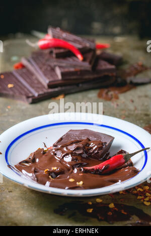 Weißen Teller mit dunklen Schokolade schmelzen und Zerkleinern von Schokolade mit frische und trockene rote scharfe Chilischoten über alte Metall Hintergrund Stockfoto