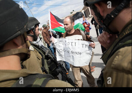 Eine palästinensische Aktivist mit einem Schild mit der Aufschrift, "Boykott die Besetzung", blickt israelische Soldaten während einer Protestaktion in der West Bank. Stockfoto