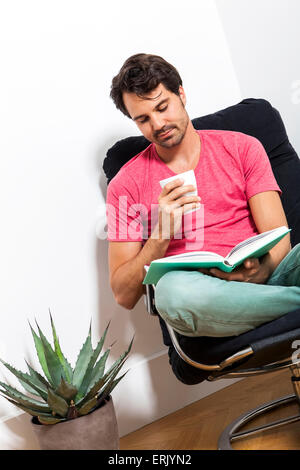 Junger Mann in Freizeitkleidung auf schwarzen Stuhl sitzend und ein Buch lesen und hält ein Glas des Getränks. Stockfoto