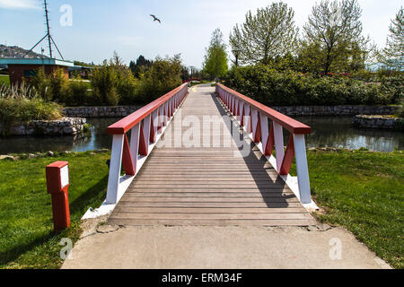 Vorderansicht der Holzbrücke mit roten und weißen Geländer am Fluss unter üppigen Feld. Stockfoto