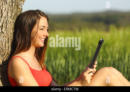 Mädchen lesen, ein Ebook oder Tablet in einem grünen Feld in einem Baum gelehnt sitzt Stockfoto