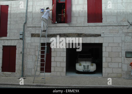 Ein Mensch malt ein Haus Rollläden in Frankreich mit einem französischen Oldtimer Citroen 2CV in der Garage unter ihm Stockfoto