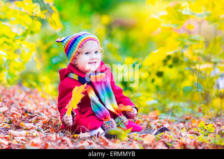 Wunderschönes Lachen Kleinkind Mädchen in einem roten Mantel und bunt gestrickte Mütze und Schal mit gelben Ahornblätter spielen Stockfoto