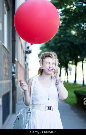 Lockige blonde Mädchen auf ihrem Handy mit großen roten Ballon neben ihr sprechen. Lächeln auf den Lippen während der Kommunikation mit ihren Freunden online. Stockfoto