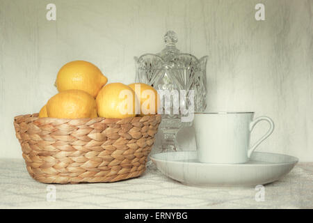 Eine Teetasse sitzt auf eine hübsche Tischdecke mit eine Kristallschüssel und einen Korb mit frischen Zitronen. Stockfoto