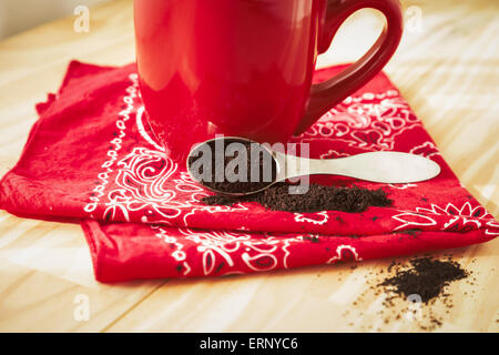 Eine rote Kaffeetasse sitzt auf einer Serviette rotes Halstuch.  Eine Kugel Kaffeesatz um verschüttet und setzt neben der Tasse. Stockfoto