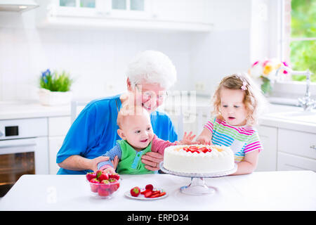 Glücklich senior Lady, liebevolle Großmutter, Backen einen hausgemachte Erdbeerkuchen mit zwei Kindern in eine weiße moderne Küche mit Fenster Stockfoto