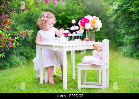 Entzückende Kleinkind Mädchen mit dem lockigen Haar trägt ein buntes Kleid an ihrem Geburtstag spielen Teeparty mit einer Puppe Teddybär Stockfoto