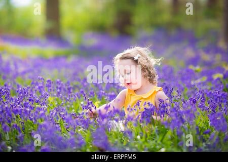 Entzückende Kleinkind Mädchen mit dem lockigen Haar trägt ein gelbes Kleid mit lila Bluebell Blumen in einem sonnigen Frühlingstagen Wald spielen Stockfoto